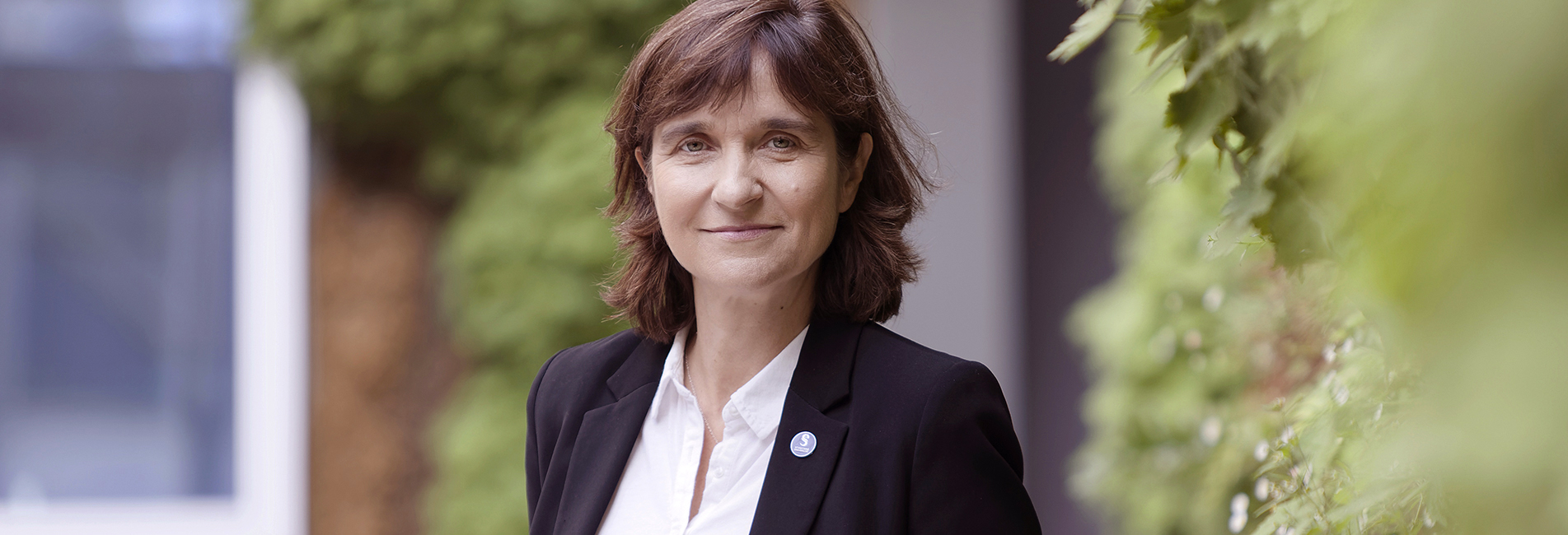 Nathalie Drach-Temam, Présidente de Sorbonne Université, elle sourit, de face sur un fond de nature vert avec une chemise blanche et une veste noire.